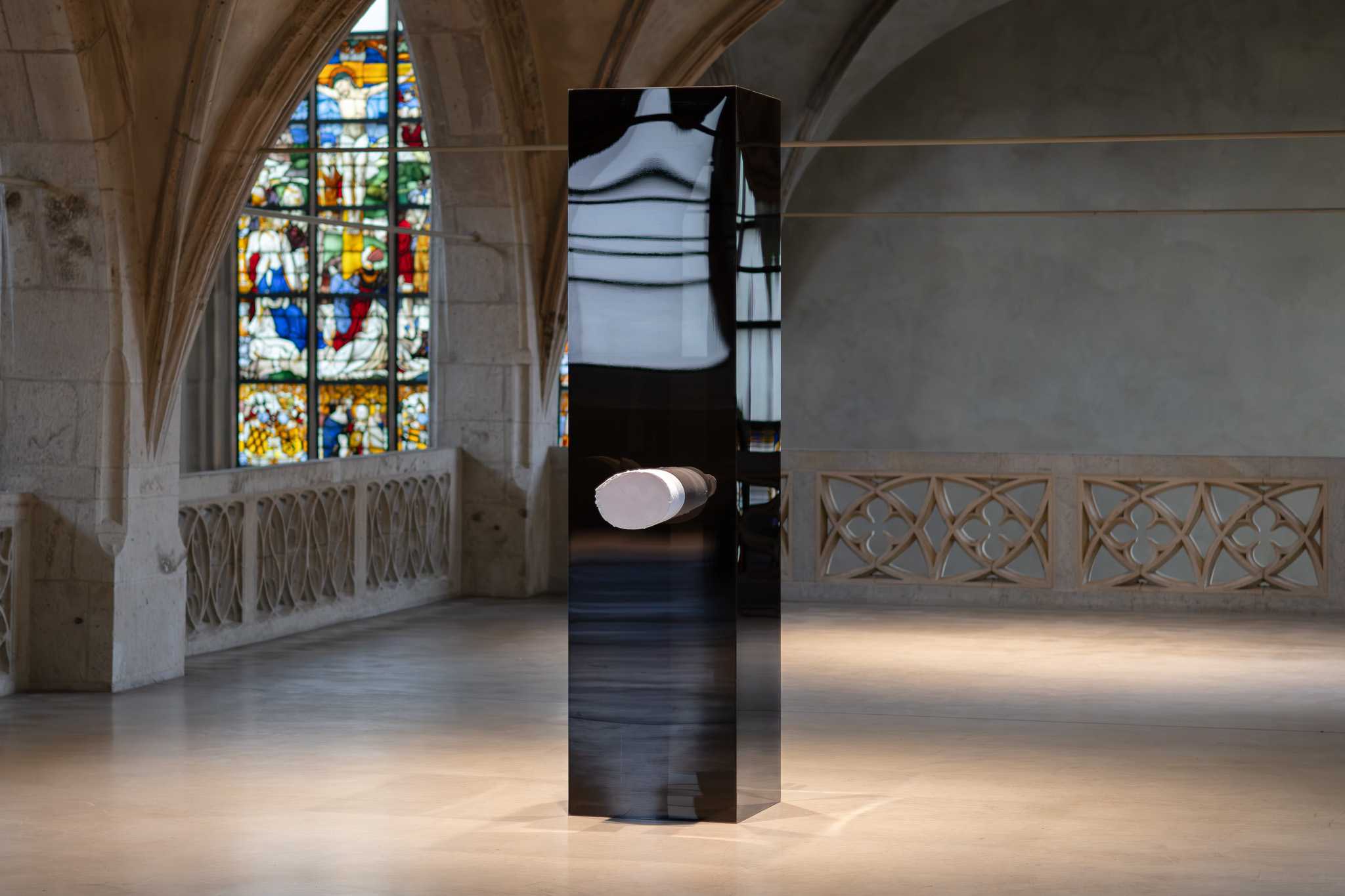 Eine hohe schwarze Stele in einem gewölbten Kirchenraum, dahinter ein Buntglasfenster; ein weißes 3D-gedrucktes Armreliquiar ragt aus ihrer Vorderseite.