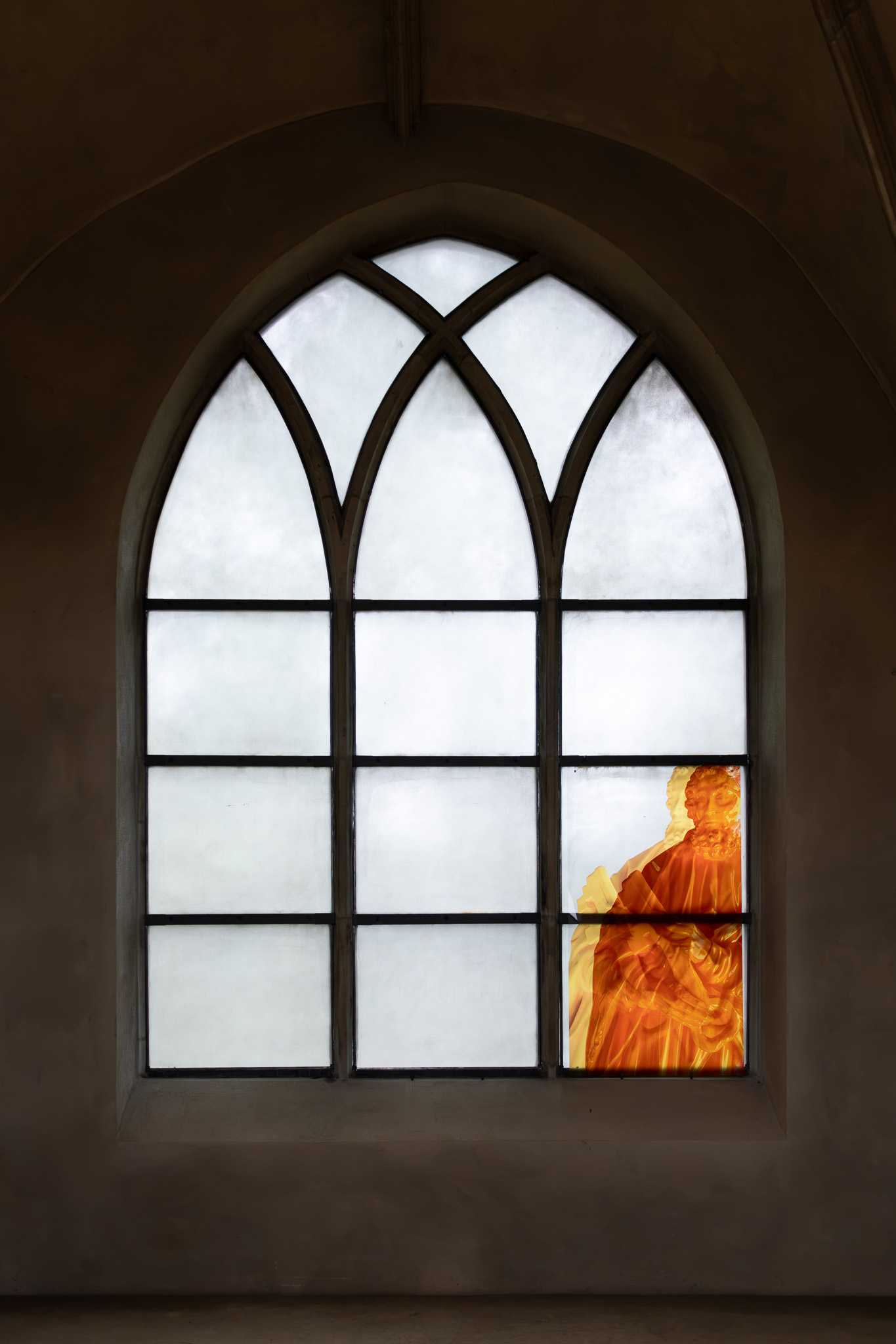 Ein tief sitzendes Spitzbogenfenster mit drei weiß verglasten Bahnen; ein durchscheinender orangefarbener Druck von Sankt Peter mit seinem Schlüssel füllt die unteren Felder der rechten Bahn.