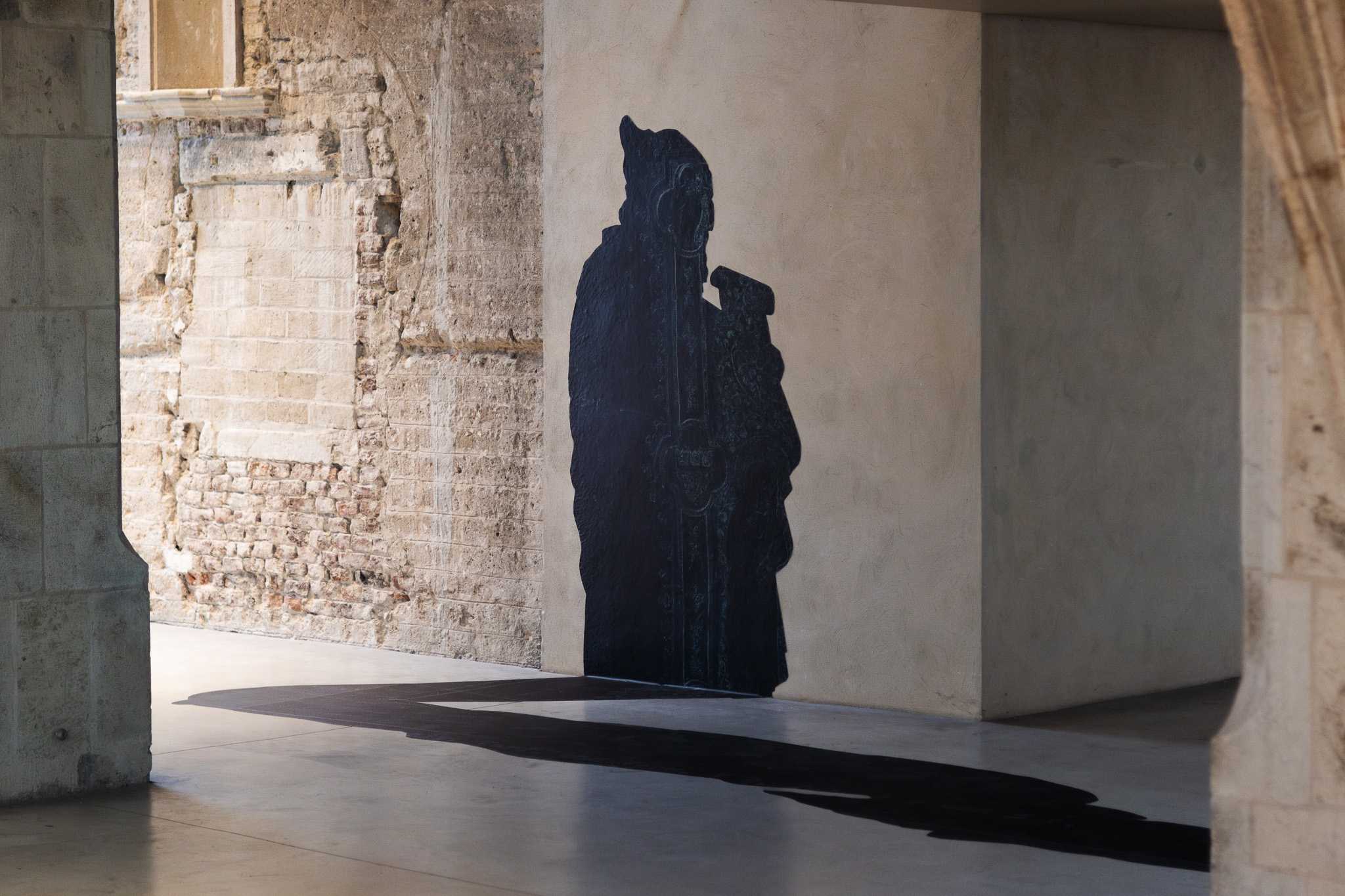 Eine schwarze Silhouette eines der Kölner Rathauspropheten mit einer Schriftrolle, angebracht an einem verputzten Pfeiler; ihr flacher Schatten erstreckt sich über den polierten Boden.