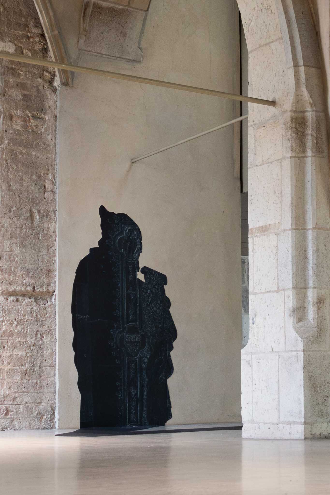 Der Prophet in voller Höhe vor einer verputzten Wandfläche, links grobes Ziegelmauerwerk, rechts ein heller Steinpfeiler, darüber quert eine schmale Hängeschiene, zu seinen Füßen ein flacher Schatten.